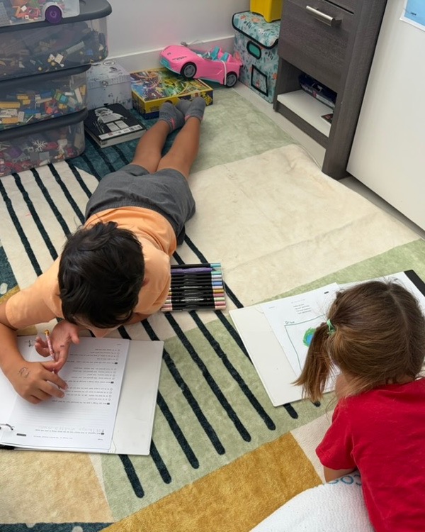 Two children lying on the floor, working through printed exercises with markers